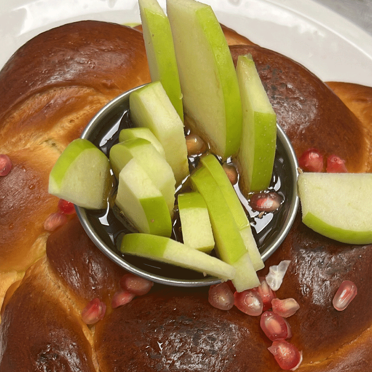 Challah bread roll with sliced apple, pomegranate seeds, and honey dipping sauce on a plate